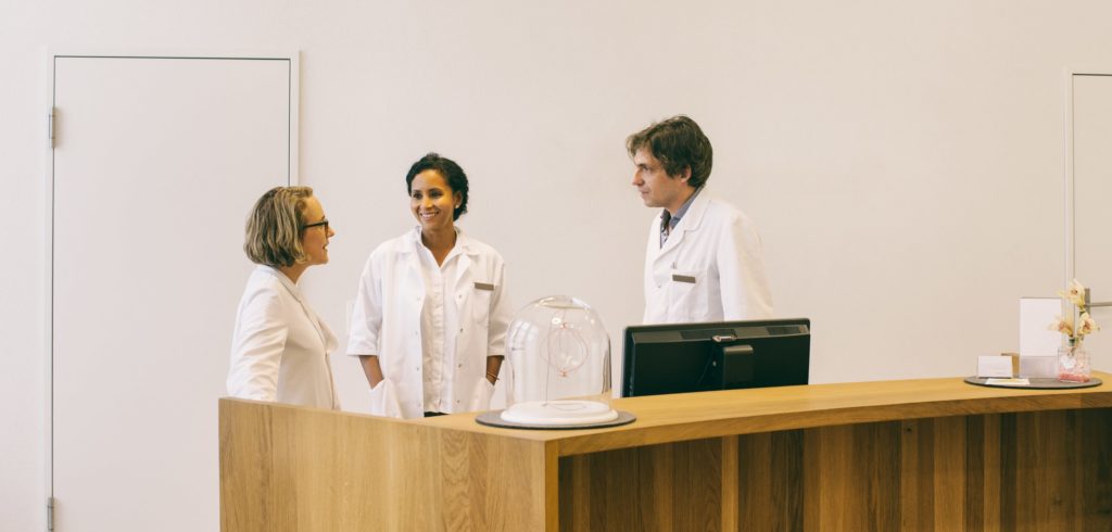 Three medical professionals in white coats standing behind a desk talking